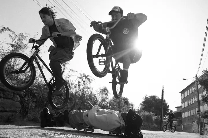 Dos ciclistas de BMX saltando sobre una persona tumbada en un monopatín, a contraluz y en blanco y negro