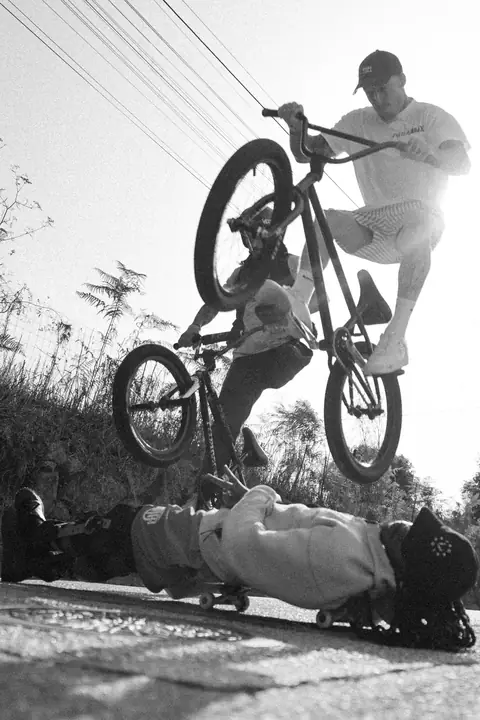 Ciclista de BMX en el aire con otro ciclista detrás, sobre un patinador tumbado, en blanco y negro