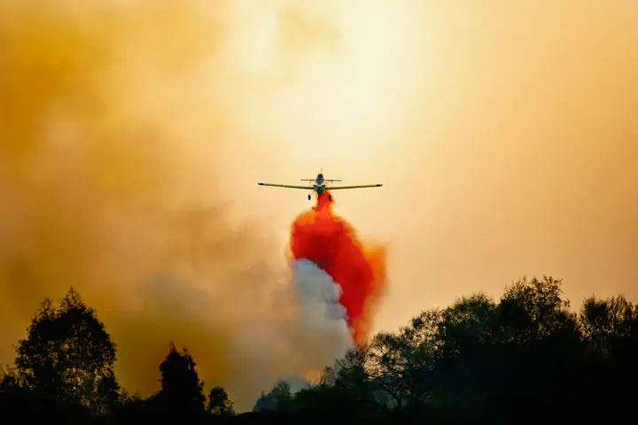 Avioneta lanzando retardante rojo sobre un incendio, entre humo anaranjado