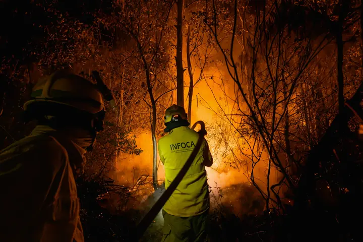 Bombero forestal dirigiendo una manguera contra las llamas de noche, con otro bombero en primer plano