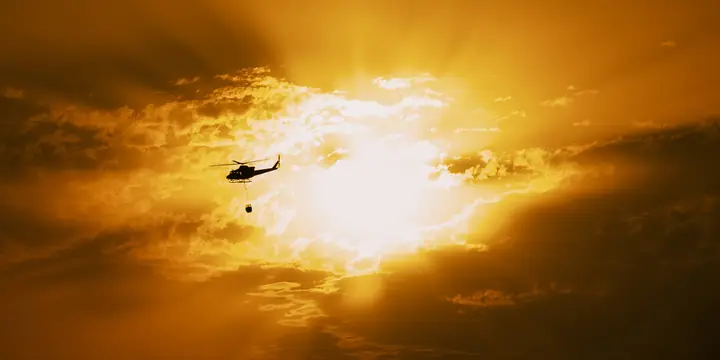 Helicóptero con helibucket a contraluz del sol entre nubes anaranjadas