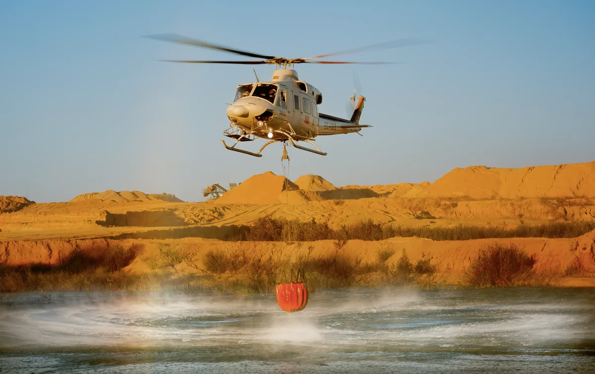Helicóptero Bell 412 cargando agua con un Bambi Bucket en una balsa de Huelva, con tierra anaranjada al fondo